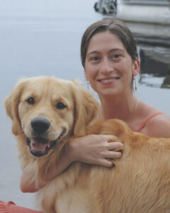 A woman sits with a dog next to water.